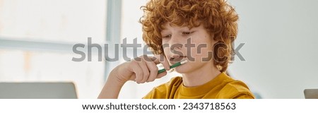 Redhead schoolboy in casual clothes holding pencil near laptop in classroom, banner