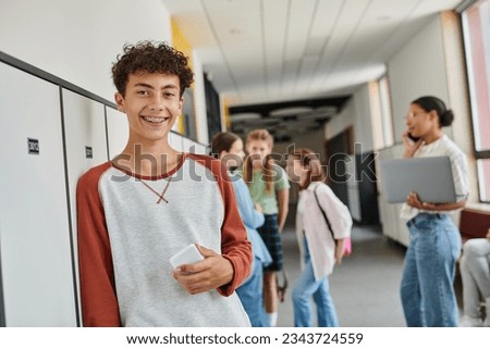 happy boy with braces holding smartphone and looking at camera during break in school hallway Royalty-Free Stock Photo #2343724559