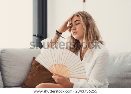 Overheated female sitting on couch in living room at hot summer weather day feeling discomfort suffers from heat waving white fan to cool herself, girl sweating dwelling without air conditioner  Royalty-Free Stock Photo #2343736255