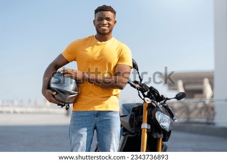 Smiling confident african american man, biker holding helmet and looking at camera near sport motorcycle. Handsome model wearing orange t shirt posing for pictures 