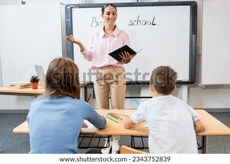 Cheerful Latinic female teacher explaining task to students. Woman stands in front of the children near the blackboard, holds a notebook and explains the material. Children listen carefully Royalty-Free Stock Photo #2343752839