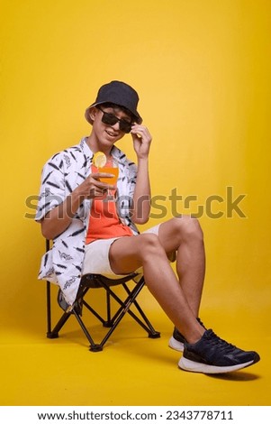 Full length of Asian guy relaxing summer holiday enjoying glass of orange juice over yellow background