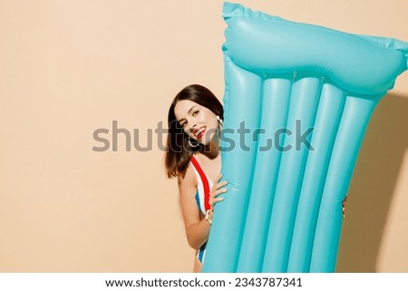 Young woman wear one-piece swimsuit straw hat lying hold blue inflatable mattress near hotel pool look camera isolated on plain pastel light beige background. Summer vacation sea rest sun tan concept