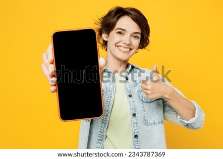 Young happy woman wear green t-shirt denim shirt casual clothes hold in hand use close up mobile cell phone with blank screen workspace area show thumb up isolated on plain yellow background studio