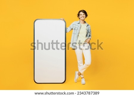 Full body smiling cheerful young woman she wear green t-shirt denim shirt casual clothes big huge blank screen mobile cell phone smartphone with area look camera isolated on plain yellow background