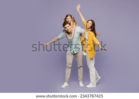 Full body young parents mom dad with child kid daughter girl 6 years old wear blue yellow casual clothes giving piggyback ride to joyful, sit on back spread hands isolated on plain purple background