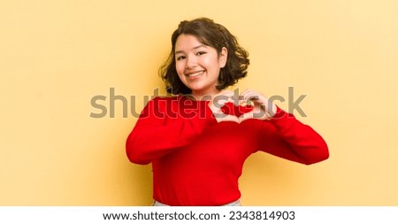 pretty hispanic woman smiling and feeling happy, cute, romantic and in love, making heart shape with both hands