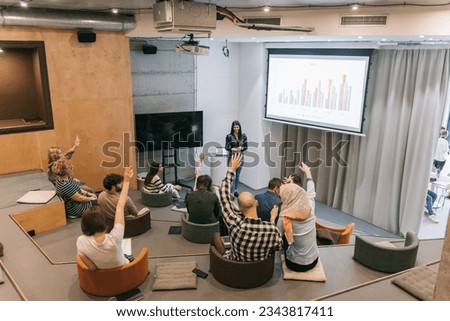 Young and beautiful business woman presenting a sales graph to a diverse group of people in a small lecture hall, discussing with them, and having a vote Royalty-Free Stock Photo #2343817411