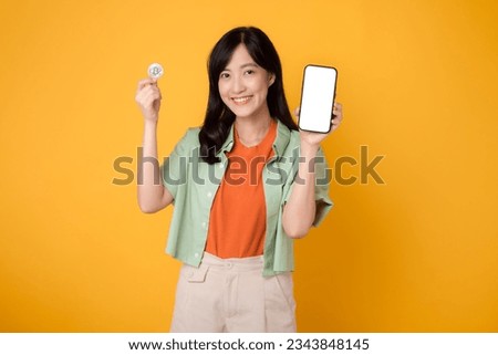 future finance with a cheerful young Asian woman 30s, donning orange shirt and green jumper, showcasing smartphone screen display and crypto currency coin on yellow background.