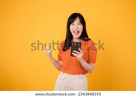 Happy Asian woman 30s, wearing orange shirt, using smartphone with fist up hand sign on vibrant yellow background. Exciting new app concept. Royalty-Free Stock Photo #2343848193