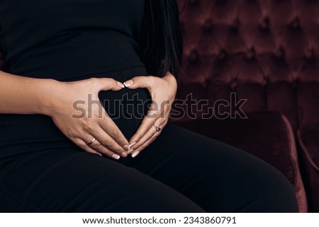 Cropped image pregnant woman strokes hugging the belly abdomen enjoying pregnancy showing heary sign symbol, sitting on maroon couch dressed black suit. Future family

