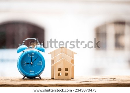 Home model and alarm clock put on the wood on sunlight building background, Business investment loan for real estate or save money for buy new house concept.