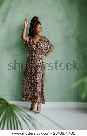 The full-body portrait of the charming black woman looking away and smiling with her right hand raised against a minty green wall. Beautiful female model. Royalty-Free Stock Photo #2343879401