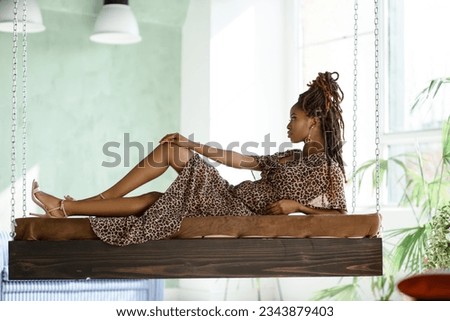 Black fashion model lying on the swing with chains in the light room with large windows. The profile full-body picture of pretty African woman. Indoor shot of girl posing. Sophisticated
 look.