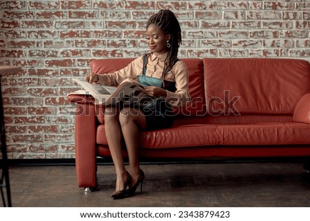 An attractive African woman sitting on the red sofa and reading good news in a newspaper against a brick background wall with copy space. Content writer, sub-editor, relationship manager, reporter Royalty-Free Stock Photo #2343879423