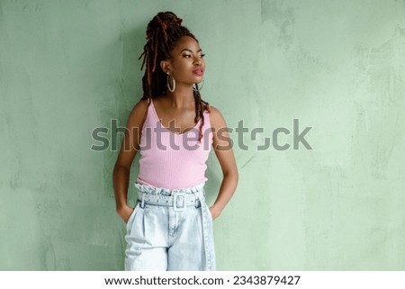 Portrait of gorgeous African girl with dreadlocks hairstyle wearing wide-leg jeans and pink top, looking away, isolated on light green wall with copy space. Beautiful confident female model. Royalty-Free Stock Photo #2343879427