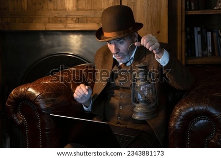 A man in a hat and a Victorian-era costume with an antique lamp in his hand looks at the laptop in a retro style. The theme is modern technology and the last century. Royalty-Free Stock Photo #2343881173