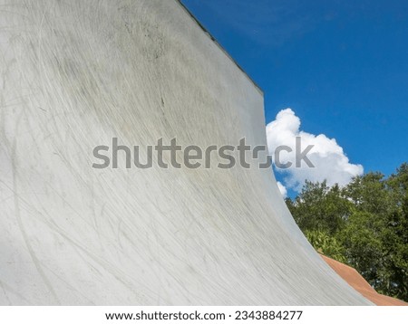 Low angle view of the steep, curved transition of a concrete vert ramp for experienced riders to perform aerial flips and spins in a public skate park, with partial view of trees, clouds, and sky