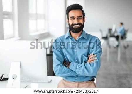 Successful young indian businessman in shirt standing with folded arms near desk, looking at camera and smiling, working in office interior, copy space Royalty-Free Stock Photo #2343892317