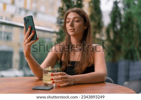 a young smiling girl sits on a summer terrace in a club bar with a cocktail and takes picture of it with her phone camera