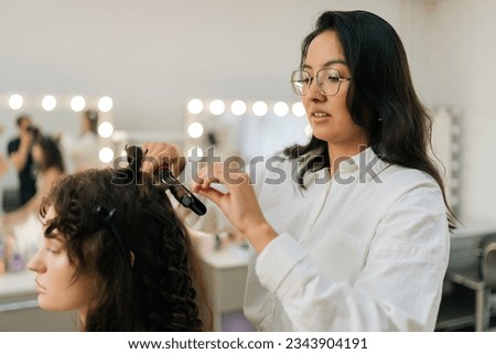 Side view of hairstylist making waves curling on woman hair. Closeup of female hairstylist in beauty salon takes lock of hair and presses with hair straightener. Concept of fashion and beauty. Royalty-Free Stock Photo #2343904191