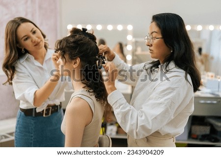 Side view of young woman being given applying highlighter for facial sculpting from female make-up artist. Hairdresser curling hair using hair iron. Concept of makeup and styling in 4 hands Royalty-Free Stock Photo #2343904209