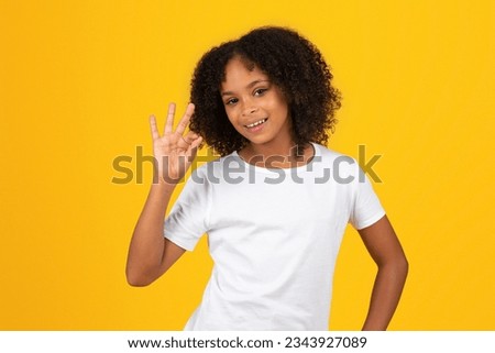Happy teenage black pupil girl in white t-shirt shows ok hand sign, isolated on yellow background, studio. Appruv study, great news, sale and back to school, ad and offer, education
