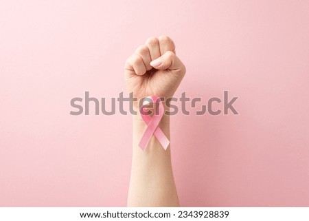Supporting Breast Cancer awareness concept. First-person perspective of strong raised hand with clenched fist. Pink ribbon attached to wrist showcased on pastel pink surface with room for text Royalty-Free Stock Photo #2343928839