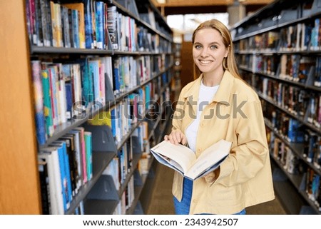 Happy pretty focused blonde girl student holding book looking at camera standing in modern university campus library or bookstore thinking of college course study reading literature, portrait.