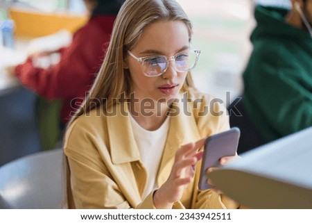 Teen girl gen z student using mobile phone looking at smartphone sitting at desk in university college campus classroom. Young blonde woman holding cellphone modern tech in university. Royalty-Free Stock Photo #2343942511