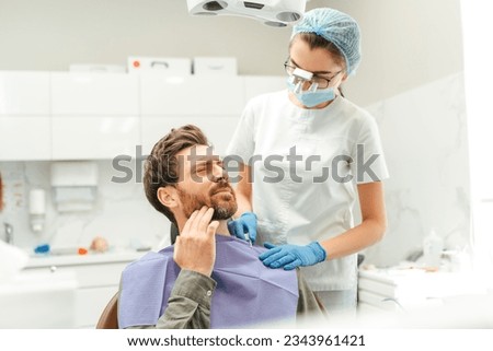 Portrait of attractive male patient sitting in dental chair, touching his cheek, toothache. Bearded man visiting dentist in modern clinic. Concept of dental treatment, health care Royalty-Free Stock Photo #2343961421