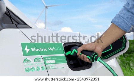 Hand insert EV charger and recharge electric car from charging station displaying futuristic battery status hologram with wind turbine farm background. Smart sustainable and alternative energy. Peruse