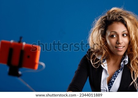 Woman with brown skin, dressed well, blonde mesh hair, nose piercing, taking a selfie with an orange smartphone and stick, seeking attractive poses for an image-focused social site