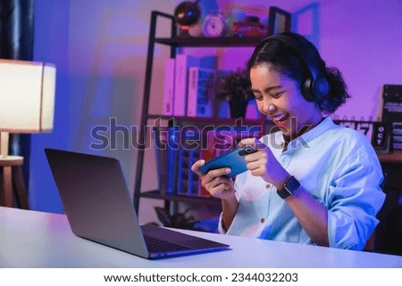 Excited Young Asian woman wearing headset and playing online game on smartphone at night modern house. Royalty-Free Stock Photo #2344032203