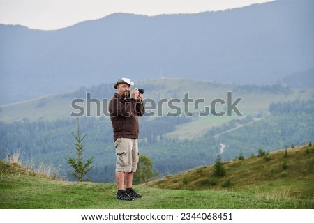 Male tourist hiking in the mountains, traveling in summer. Man standing on hill, holding camera, taking photo, admiring landscape, enjoying. Concept of tourism and adventure.