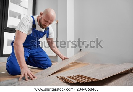 Male construction worker laying laminate wood plank on floor underlayment in living room. Bearded man in work overalls installing laminate timber flooring in apartment under renovation. Royalty-Free Stock Photo #2344068463
