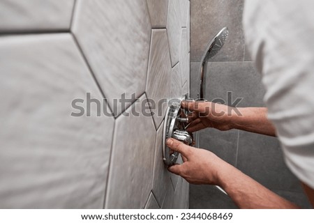 Close up of man standing by the wall with ceramic tile and installing shower faucet with metal handle in apartment. Male plumber working on bathroom renovation at home. Plumbing works concept. Royalty-Free Stock Photo #2344068469