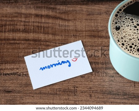 Handwritten over white card "morning" and a green coffee mug on a wooden table. Good morning message with copy space.
