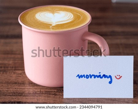 Handwritten over white card "morning" and a pink coffee mug with art heart shape on a wooden table. Good morning message with copy space.
