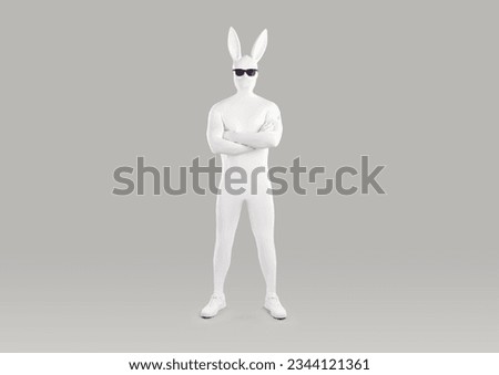 Full length portrait of faceless unrecognizable person in white spandex costume, hare mask with long ears and black glasses. Incognito funny man in bodysuit with crossed arms on grey background. Royalty-Free Stock Photo #2344121361