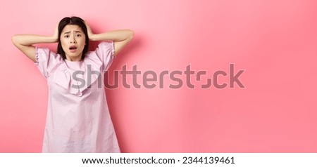 Frustrated asian teen girl panicking, holding hands on head and screaming scared, standing anxious on pink background.