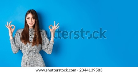 Confident adult woman in dress with natural hair color, showing okay signs and smiling, like and approve something good, standing on blue background.