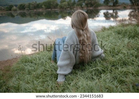back view of the blond woman dressed sweater and jeans, siting  on the grass near  the lake with beautiful landskape. The girl enjoy the nature  in her trip