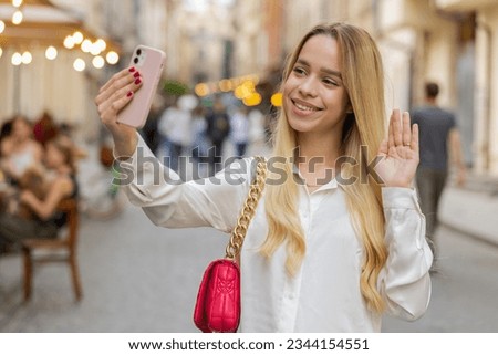 Blonde young woman blogger taking selfie on smartphone, communicating video call online with subscribers, recording stories for social media vlog outdoors. Teenager girl walking in urban city street