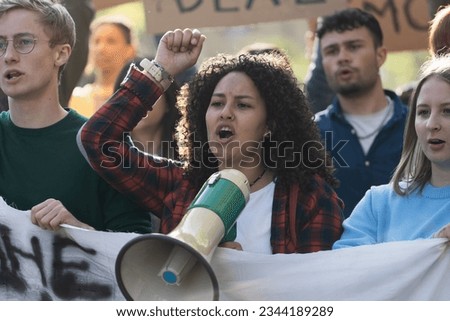 Student Activist Leading Protest with Megaphone - A close-up of a young curly-haired woman leading a generic student protest, rallying her peers with passion and a megaphone Royalty-Free Stock Photo #2344189289