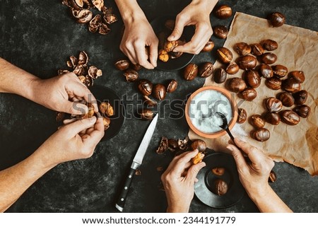 Close up of hands of three people peeling roasted chestnuts on black table background. Lifestyle family meal concept. Dark low key photo. Top view. Royalty-Free Stock Photo #2344191779