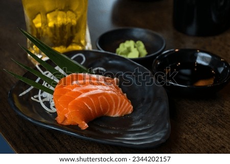 Slices of salmon sashimi on a black dish with a mug of beer, soy sauce and wasabi.