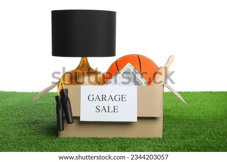 Different stuff in box with sign Garage Sale on green grass against white background