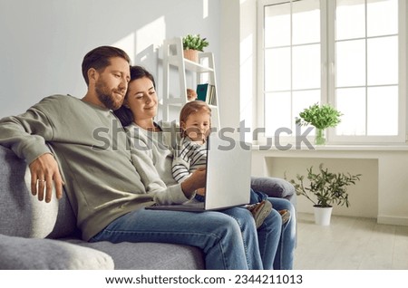 Happy young family with little child girl sitting on the sofa using modern laptop together. Smiling parents resting on couch enjoying weekend watching funny cartoon online or talking on video call.