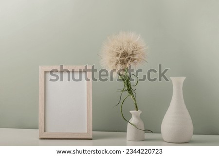 Empty photo frame and white vase with dandelion and empty vase on the table. Minimalistic interior.